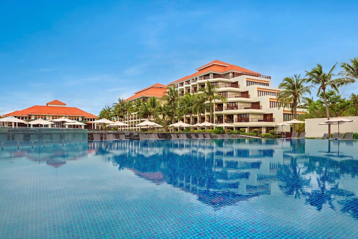 Large outdoor infinity pool at a Da Nang beachfront resort with sun loungers, palm trees, and resort buildings under blue sky