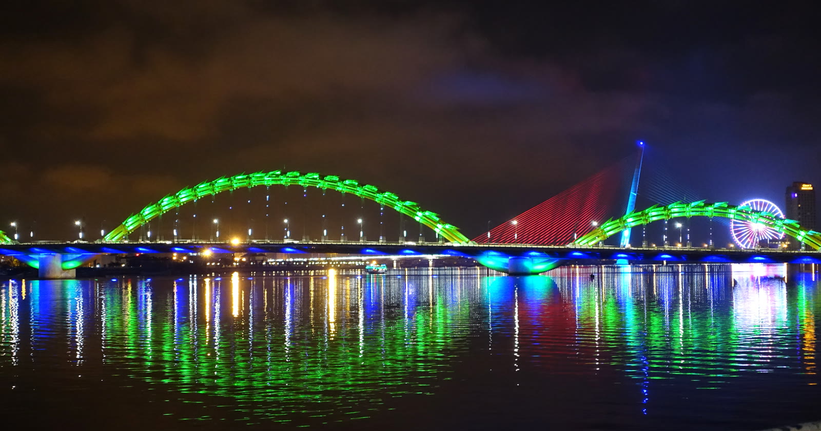 Dragon Bridge in Da Nang at night, lit blue and yellow, with the dragon head at the left of the frame and the river in front