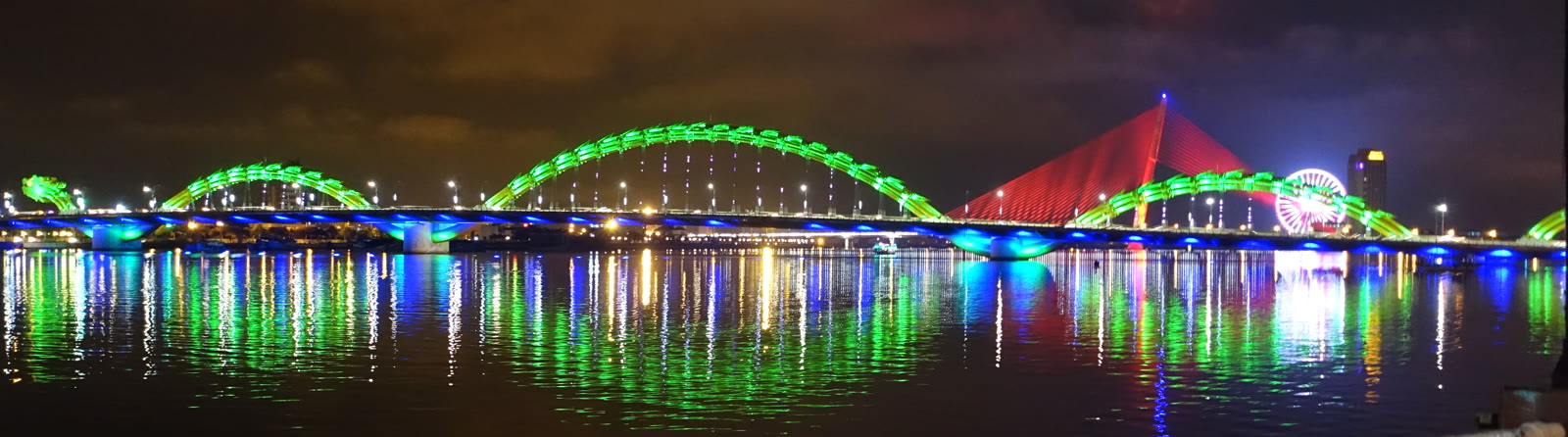 The lit span of Dragon Bridge in Da Nang at night, dragon head at the left, seen across the river